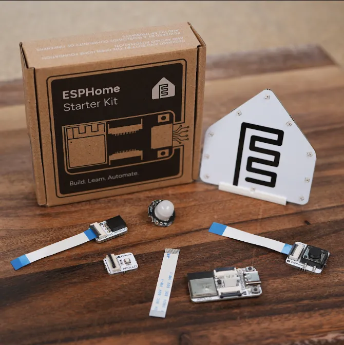 The ESPHome Starter Kit box next to its unboxed contents: notification module, main board, motion sensor, button module, ribbon cables, and stand, arranged on a wooden surface.